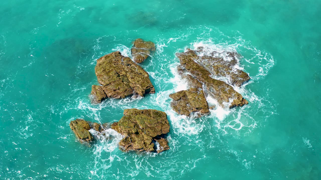 Overhead El Cardoncito frame where white surf curls around the right-side rocks and the lower rock outcrop sits in calmer turquoise water, with no visible people.