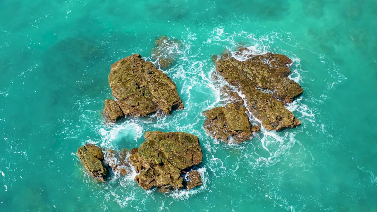 Aerial view over El Cardoncito rocks as white foam wraps around the upper and lower edges of the brown outcrops in turquoise water, no visible people.