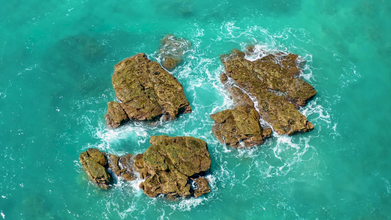 Overhead El Cardoncito frame with the rock cluster slightly right of center, open turquoise water around it, and surf breaking on the exposed stone, no visible people.