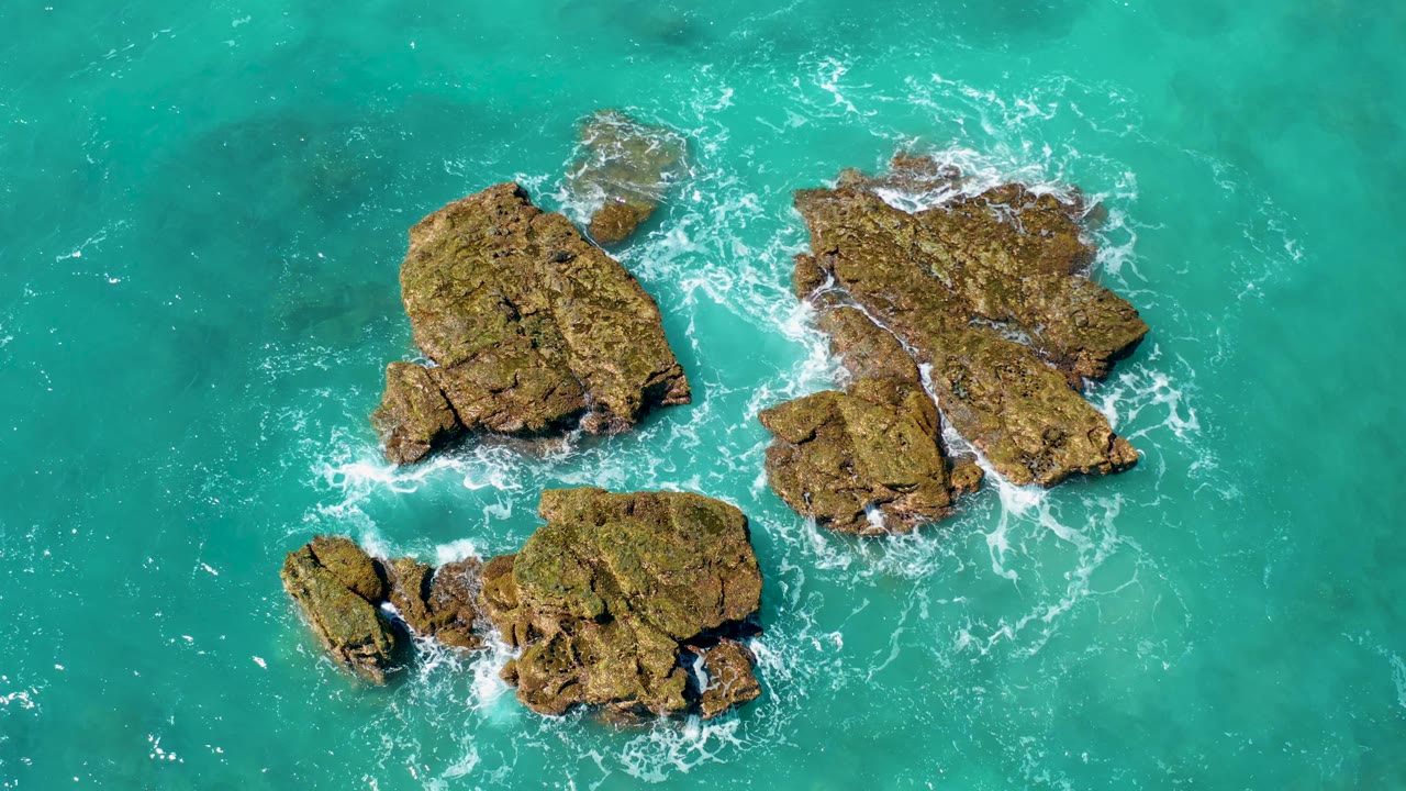 High aerial frame from El Cardoncito showing jagged brown rocks with white wave foam collecting in the gaps between the outcrops, no visible people.