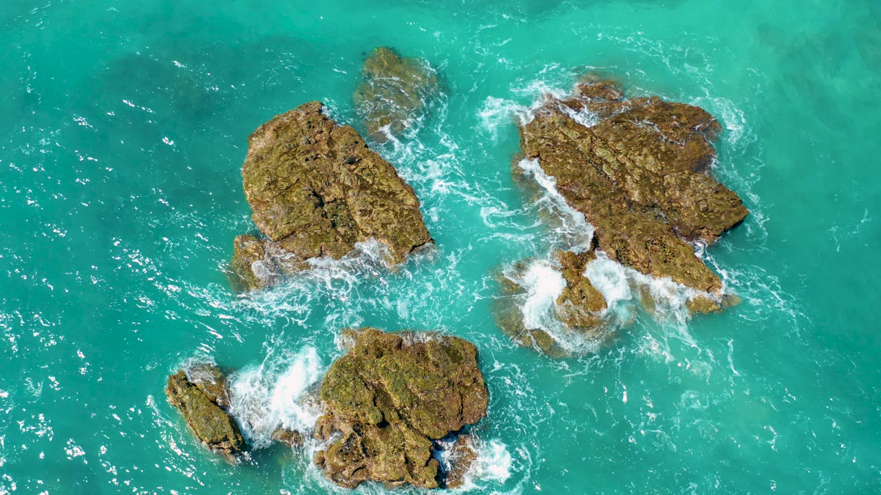 Overhead view of turquoise water around three tan rock outcrops near El Cardoncito, Baja California Sur, with white foam starting to wrap their edges and no visible people.