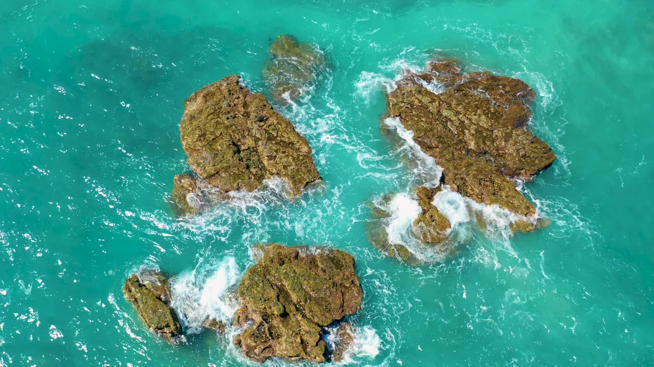 Aerial overhead frame of El Cardoncito rock outcrops in turquoise water, with white foam trailing from the lower and right rocks and no visible people.
