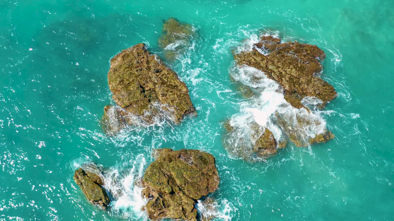Top-down view near El Cardoncito with the main rocks shifted across the frame as a larger patch of white surf breaks over the right outcrop, with no visible people.