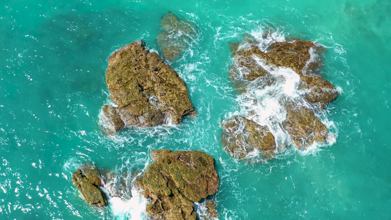 Overhead El Cardoncito coastal frame with a broad left rock, a foamy right rock, and a smaller lower rock surrounded by turquoise water, with no visible people.