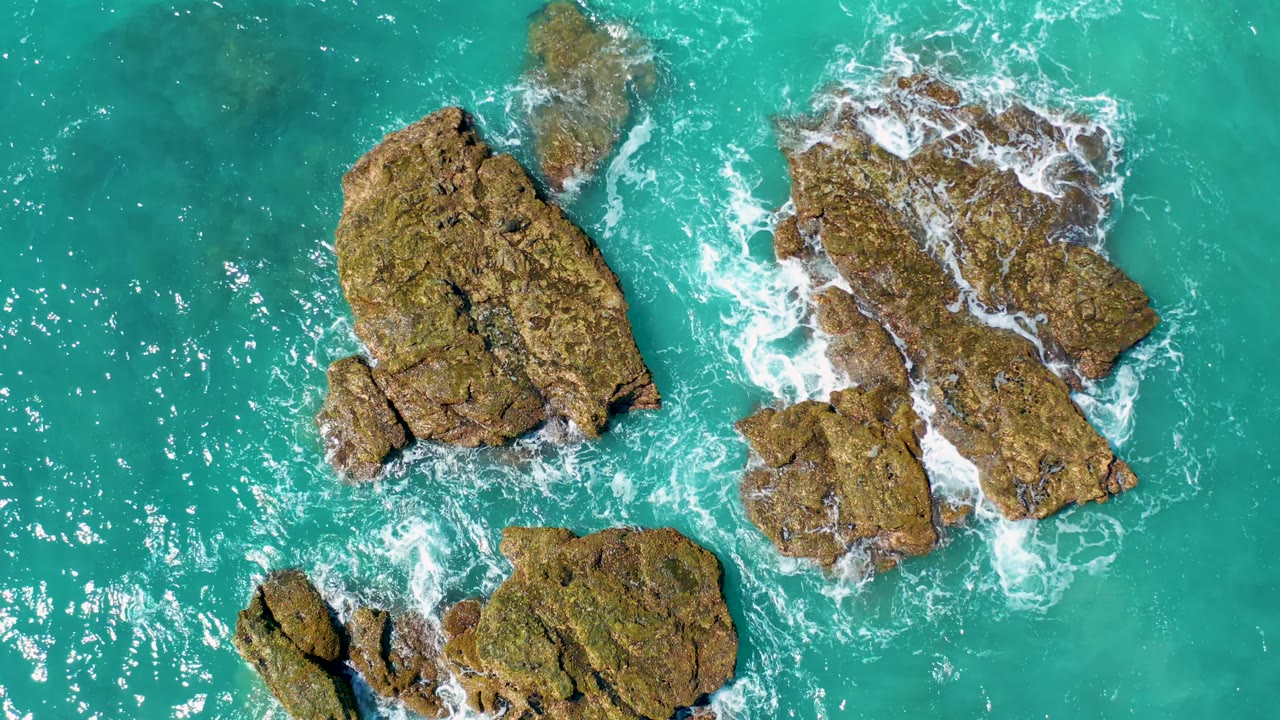 Aerial frame over El Cardoncito shows multiple uneven rock outcrops surrounded by turquoise sea, with white water collecting in narrow gaps and no visible people.