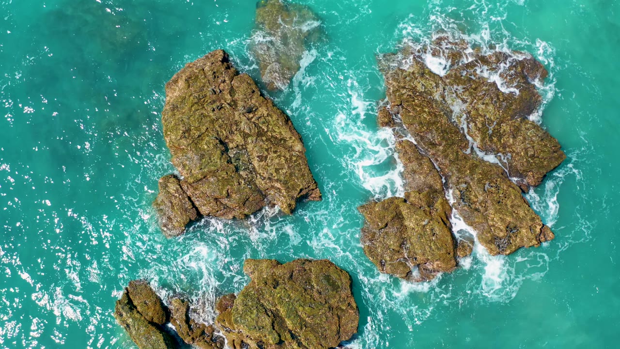 Top-down view in El Cardoncito frames the rock group slightly right of center, with bright sun reflections on the turquoise water and no visible people.