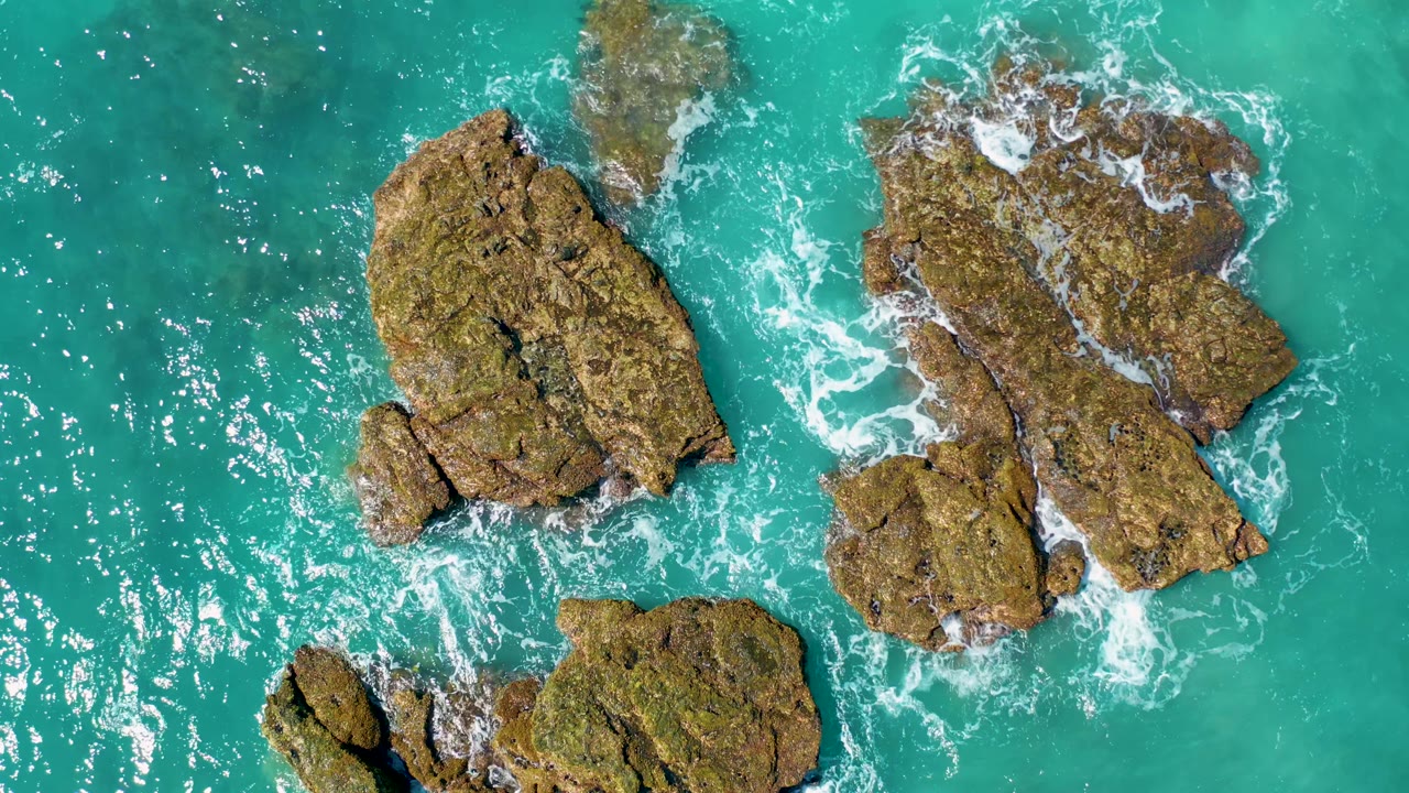 Aerial view over El Cardoncito places two large rock masses across the center, with foamy water breaking along their edges and no visible people.