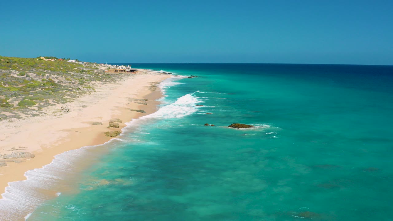 Aerial view at El Cardoncito, Baja California Sur, showing turquoise water washing onto an empty sandy beach with dunes and no visible people.