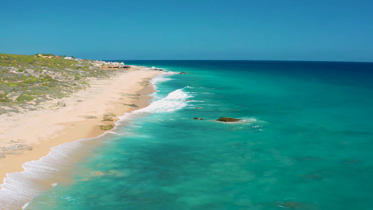 At 0:01, white surf lines run diagonally along the El Cardoncito beach while clear turquoise water fills the right side and no visible people appear.
