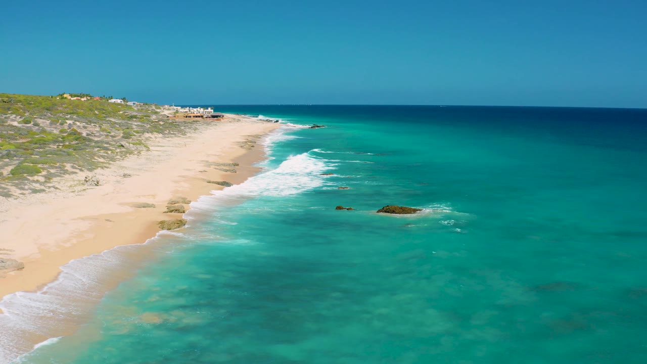 The frame shows El Cardoncito's sandy shoreline curving under bright daylight, with green-blue shallows, small rocks offshore, and no visible people.