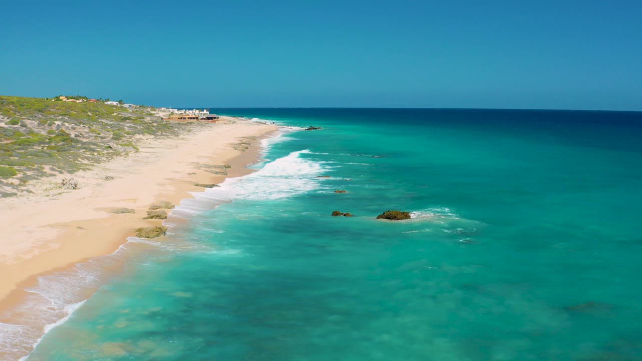 At 0:03, the aerial composition keeps the empty beach on the left and turquoise ocean on the right, with surf breaking in a narrow white band and no visible people.