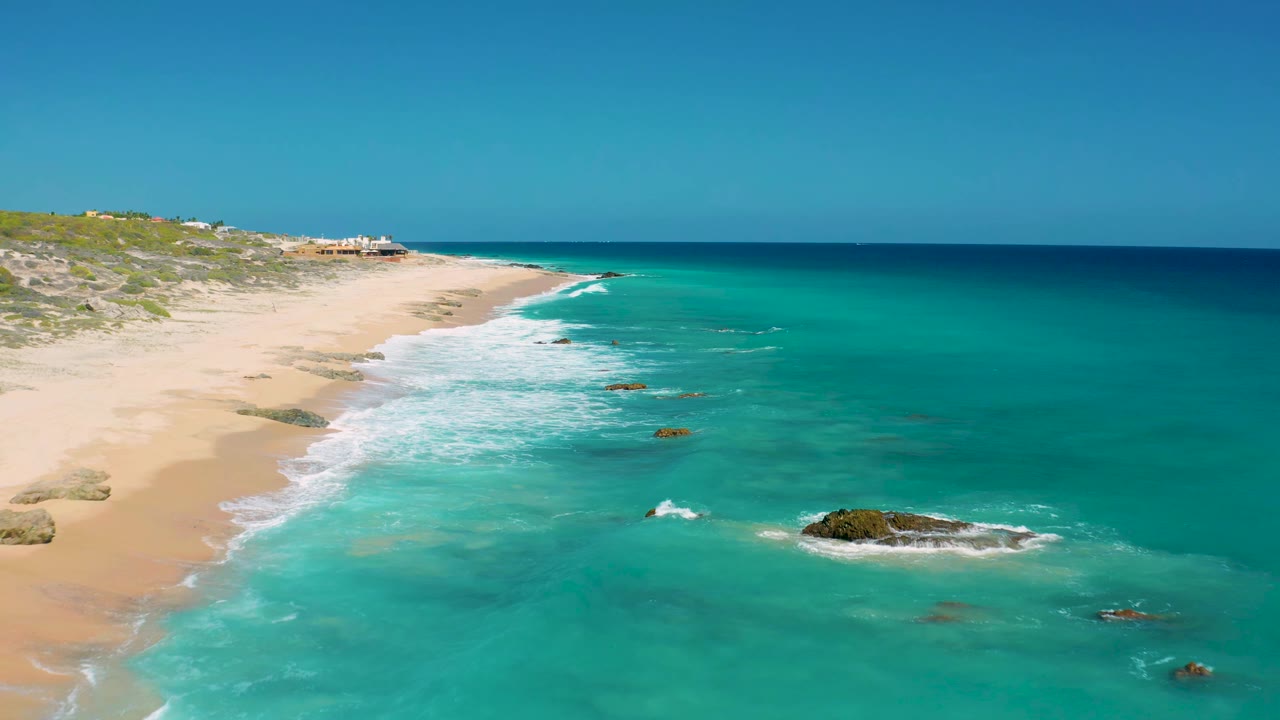 El Cardoncito aerial view with the shoreline running diagonally, white waves spreading across pale sand, scattered rocks in clear turquoise water, and no visible people.