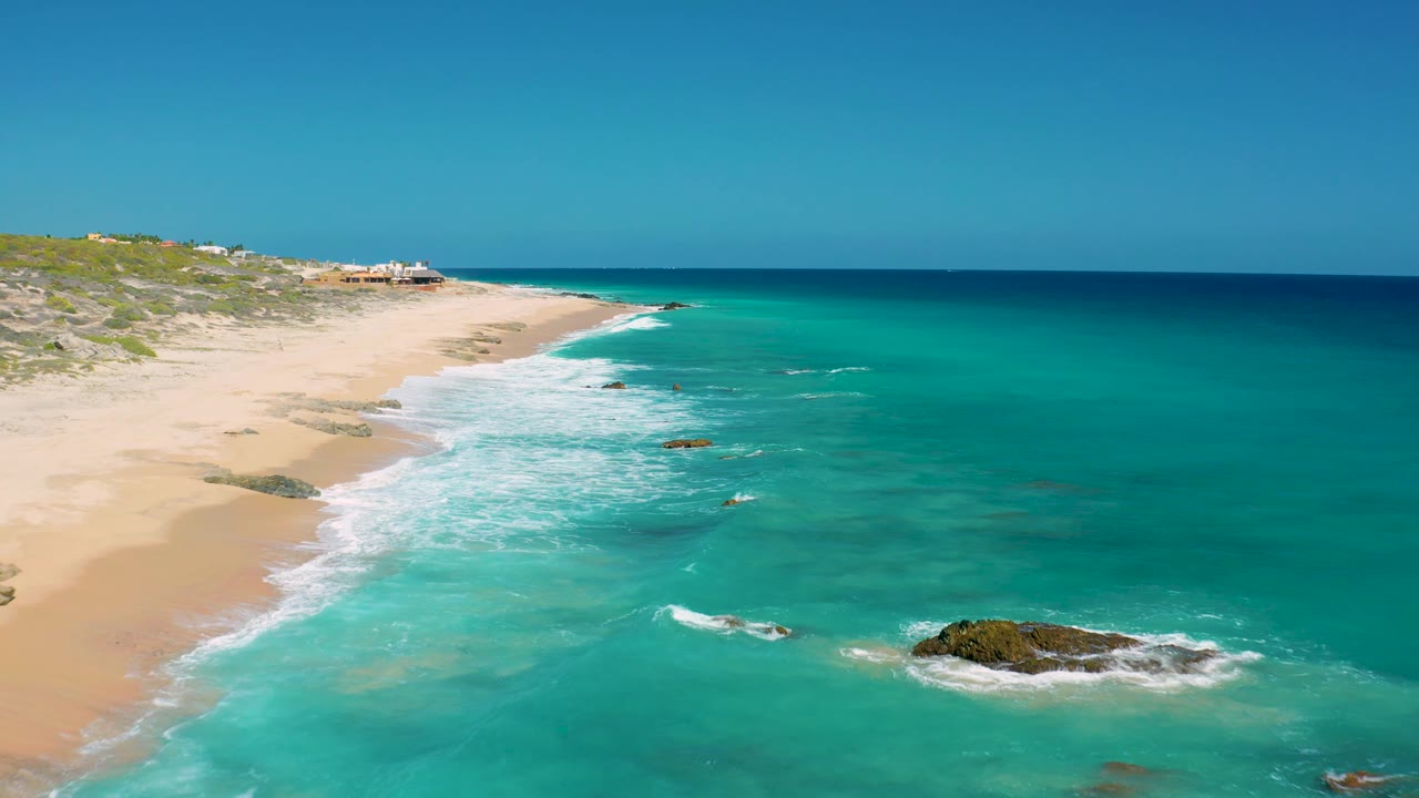 Aerial frame from El Cardoncito showing blue ocean bands, foamy surf beside the sand, a larger dark rock near the lower right water, and no visible people.