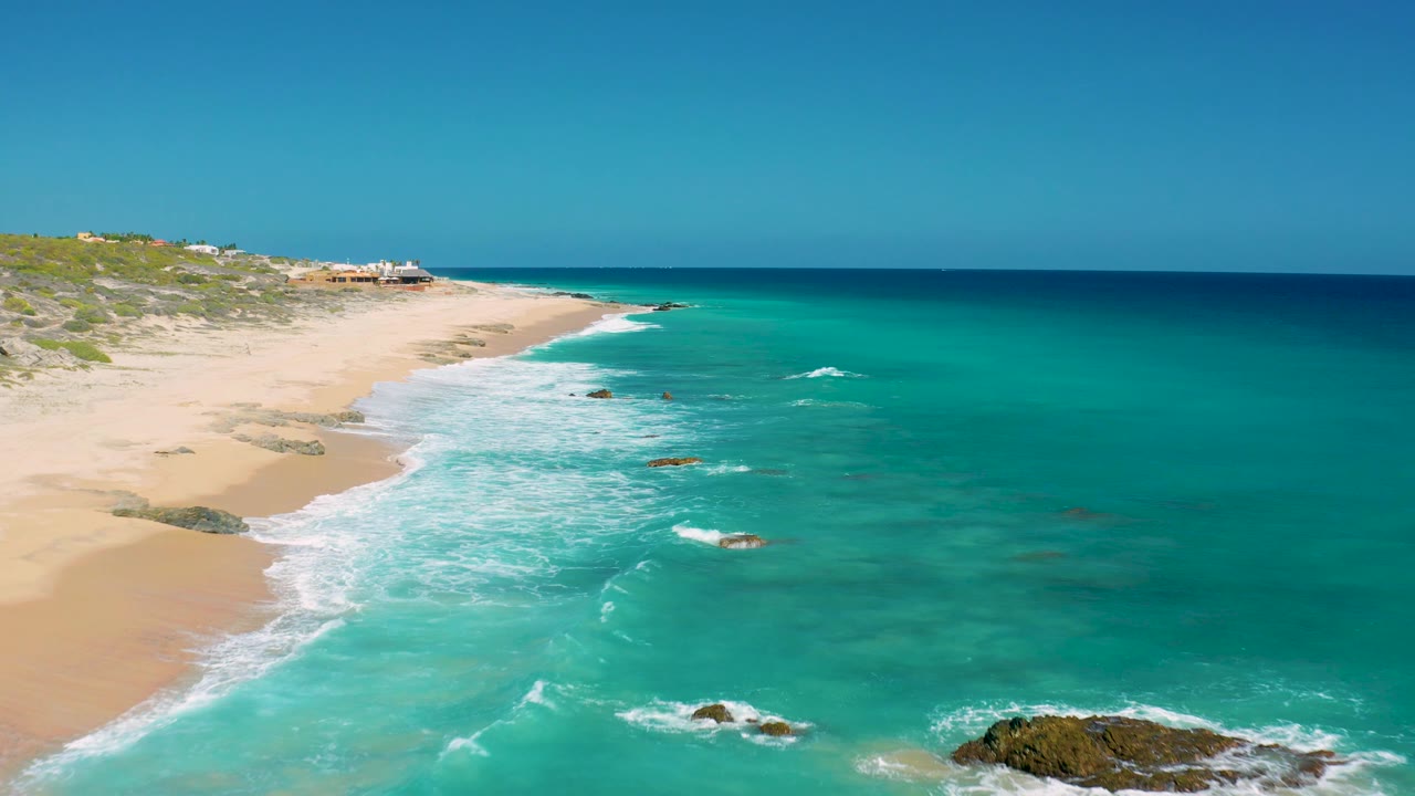 El Cardoncito beach seen from above, with foamy waves sweeping toward the sand, dark rocks in the shallows, low dunes at the upper left, and no visible people.