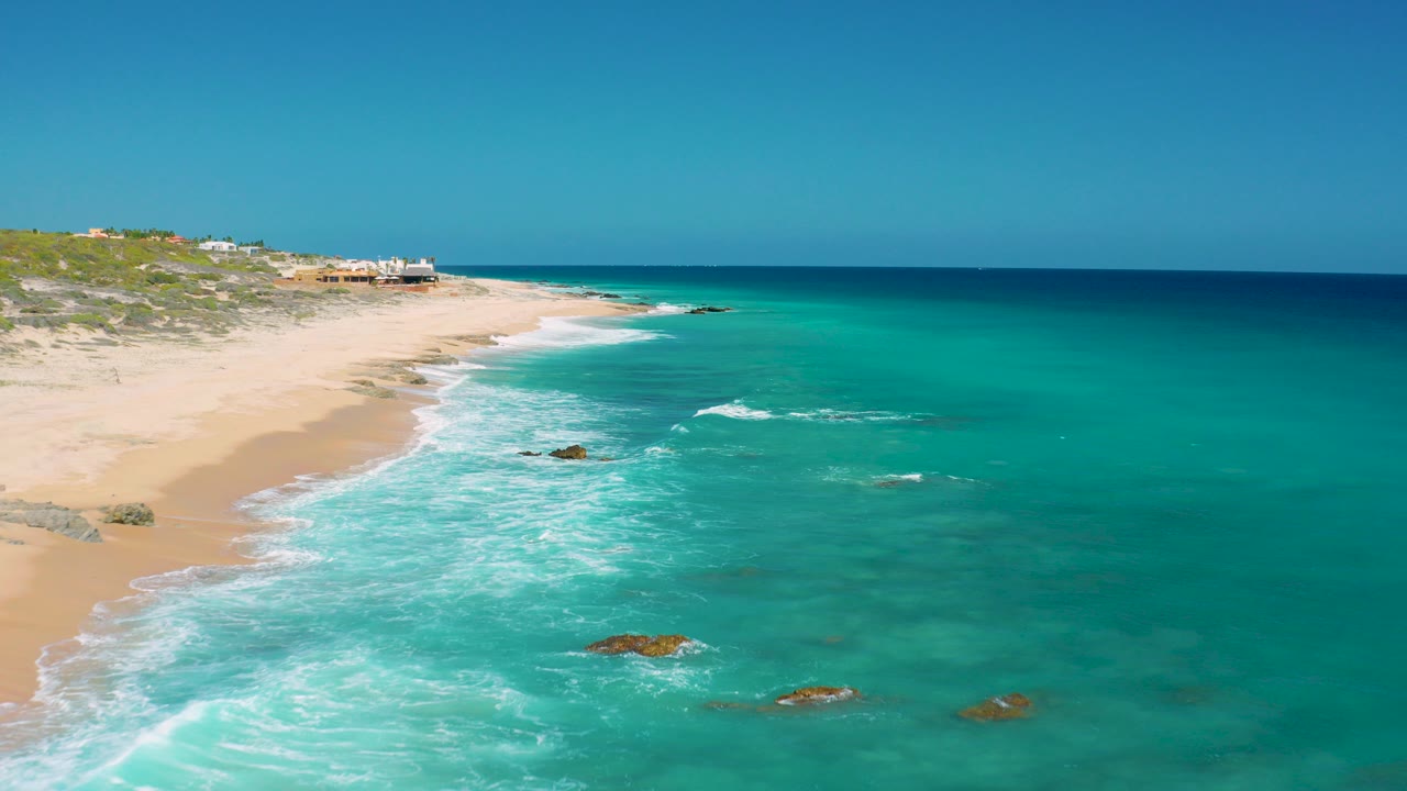 Elevated view of sandy beach, turquoise water, white surf, and rocks under a clear sky.