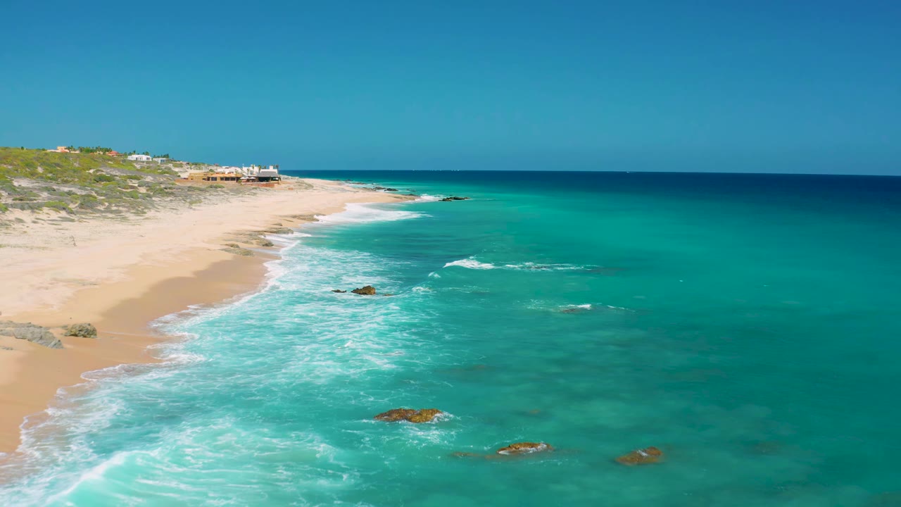 Aerial view at 0:01 over El Cardoncito showing a longer sweep of white surf along the sand, blue-green water around offshore rocks, low dunes, distant buildings, and no visible people.