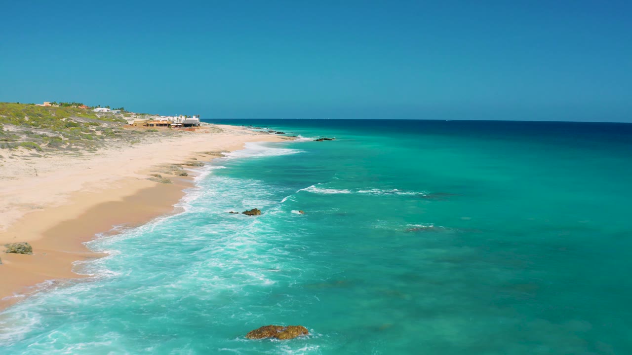 Aerial frame over El Cardoncito with the beach receding from left foreground to upper center, broad turquoise water filling the right side, white foam over shallow water, exposed rocks, and no visible people.