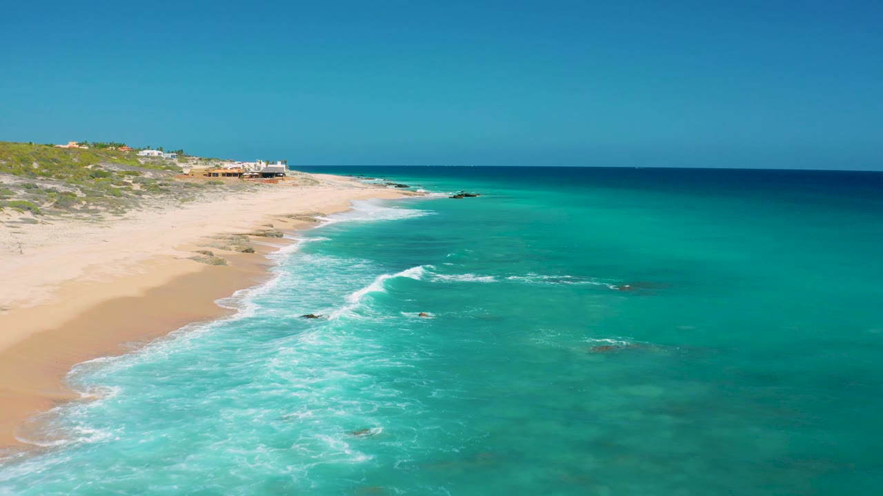 Mid-clip aerial frame at El Cardoncito where white surf cuts diagonally from the lower left toward the center, with sandy beach, low vegetation, scattered rocks, clear blue sea, and no visible people.