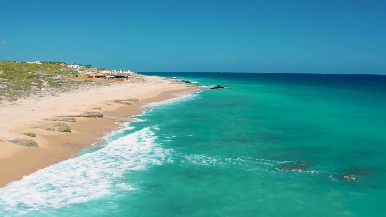 Aerial frame in El Cardoncito showing the surf line stretching diagonally along pale sand, with clear blue-green ocean and no visible people.
