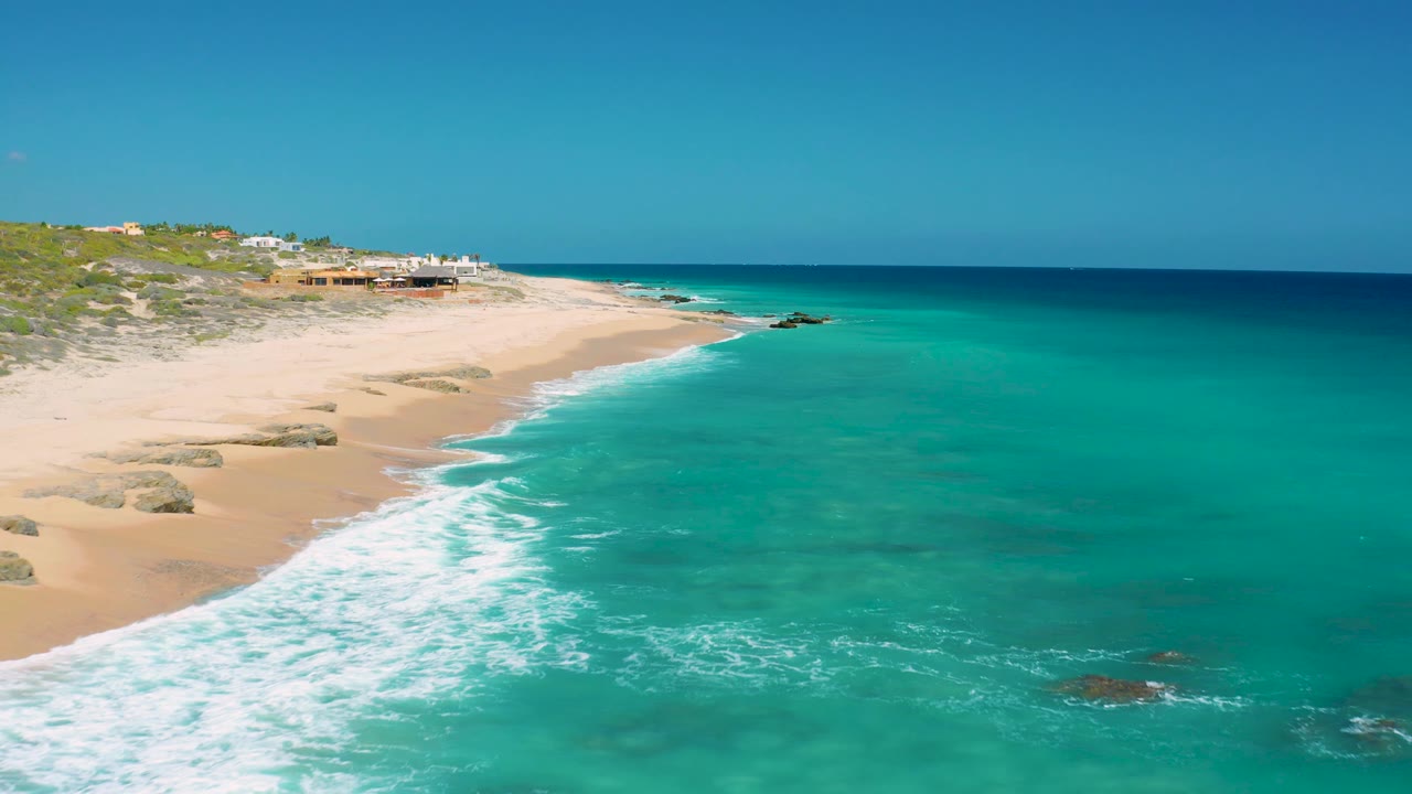Aerial frame over El Cardoncito with more open turquoise water in view, sandy shore at left, low vegetation inland, distant coastal buildings, and no visible people.