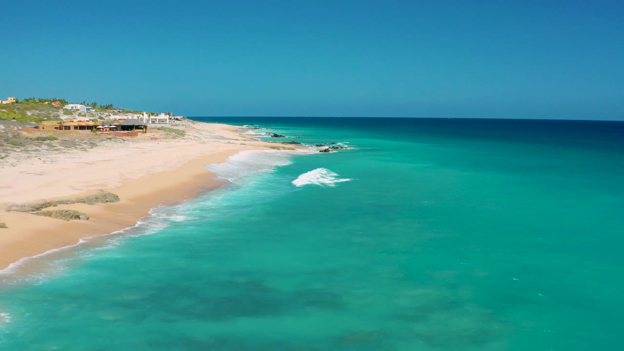 Aerial frame from El Cardoncito, Baja California Sur showing turquoise water, a sandy beach curving along the left side, low beach homes on the bluff, and no visible people.
