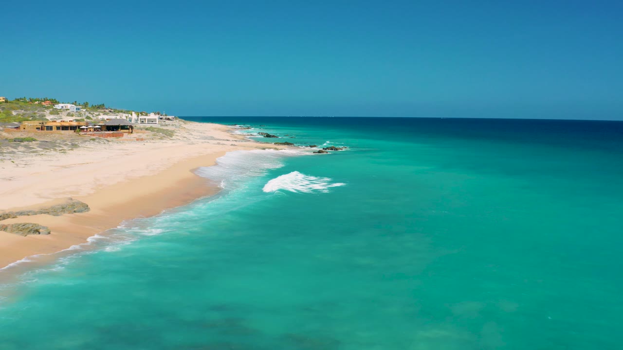 Aerial frame from El Cardoncito, Baja California Sur showing a broader stretch of turquoise ocean, white surf near the sand, beach houses above the shore, and no visible people.