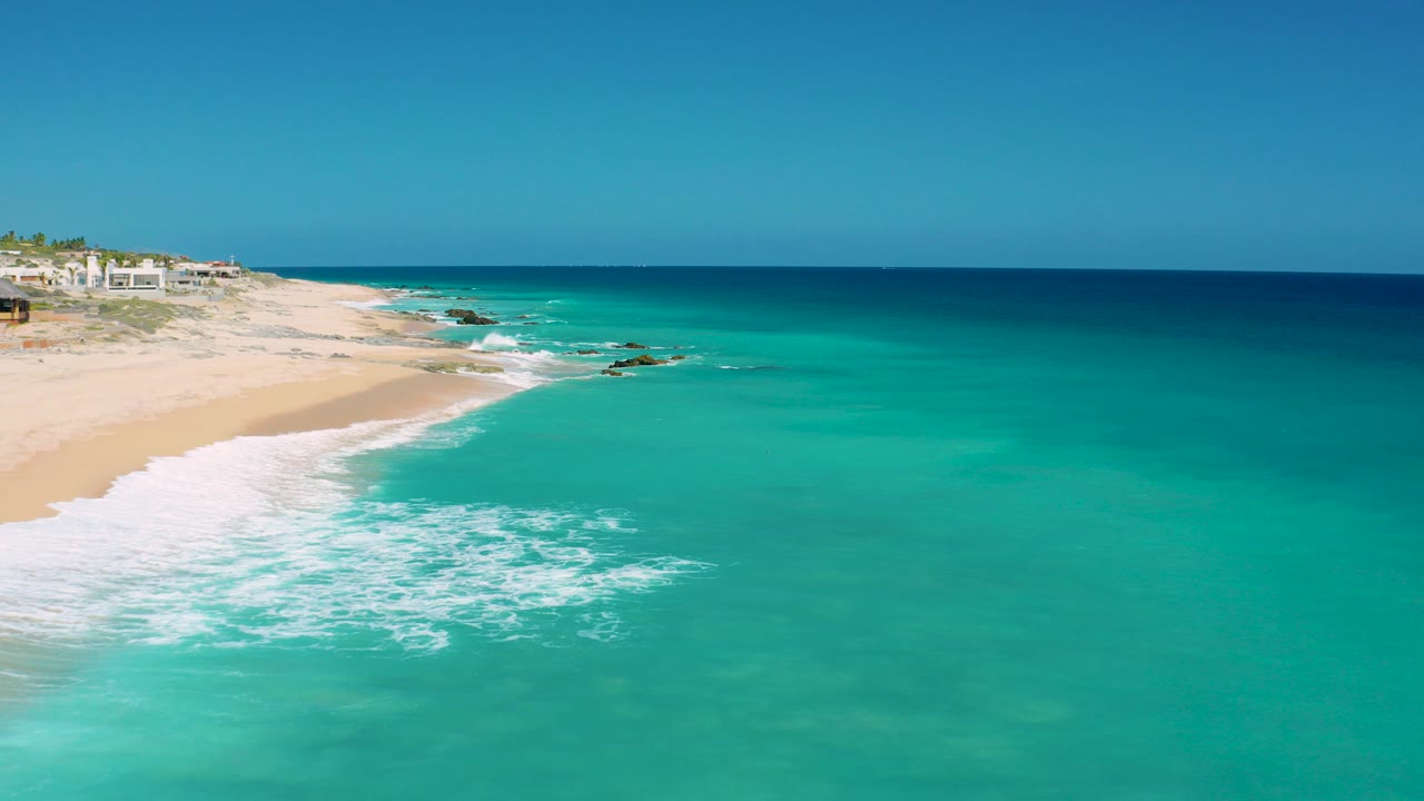 Aerial frame over El Cardoncito, Baja California Sur, with turquoise surf spreading onto pale sand, coastal buildings at upper left, dark rocks offshore, and no visible people.