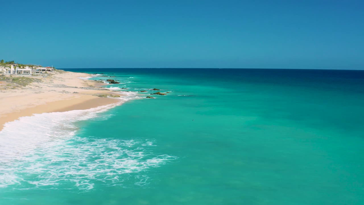 Aerial frame from El Cardoncito showing a broad turquoise shallows area, a curved strip of white surf, pale sand at left, distant rocks, and no visible people.