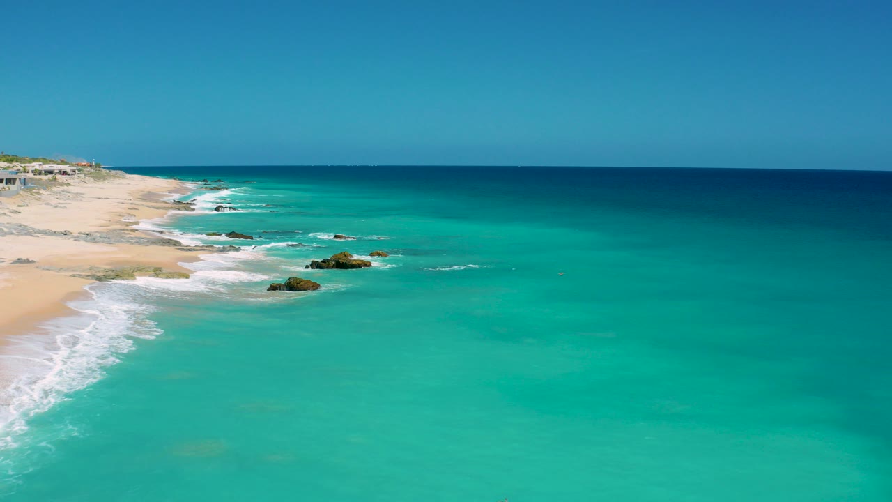 Aerial frame over El Cardoncito showing pale beach on the left, turquoise water in the foreground, deep blue horizon water, scattered rocks, and no visible people.