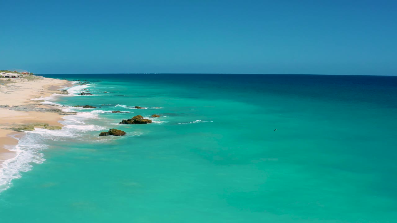Aerial frame from El Cardoncito showing more open turquoise water, the beach receding along the left side, rocks surrounded by white foam, and no visible people.