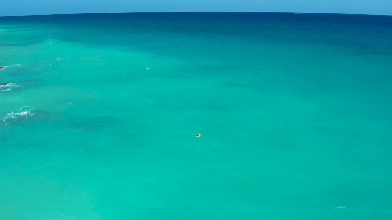 Mid-run view over clear turquoise sea near El Cardoncito, with a small object on the water, broken white wave patches to the left, and no visible people.