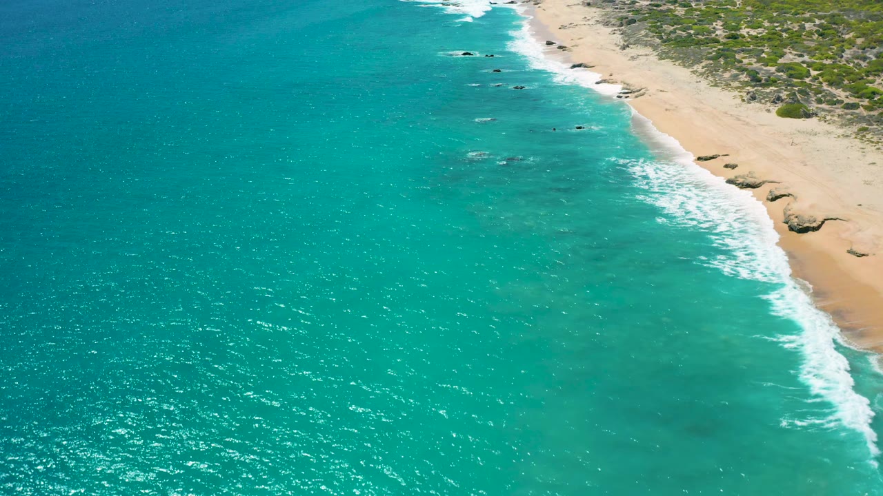 Aerial frame over El Cardoncito with open turquoise water in the foreground, a diagonal band of white surf, pale beach sand at right, scattered shoreline rocks, and no visible people.