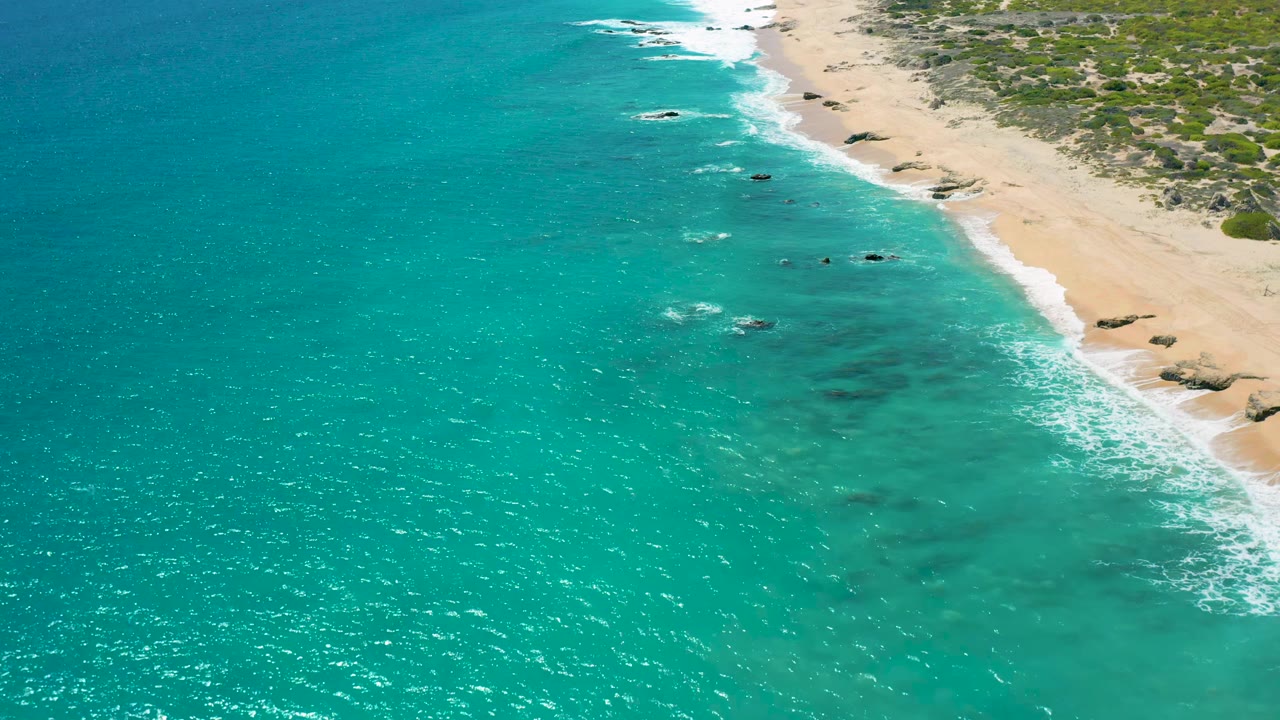 El Cardoncito coastline seen from above, with bright turquoise shallows, a narrow white surf line, pale sand, scattered dark rocks near shore, and no visible people.