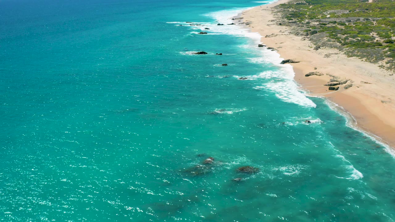 A high oblique view of El Cardoncito shows shimmering turquoise sea, a foam band meeting pale sand, several dark rocks offshore, low green vegetation inland, and no visible people.