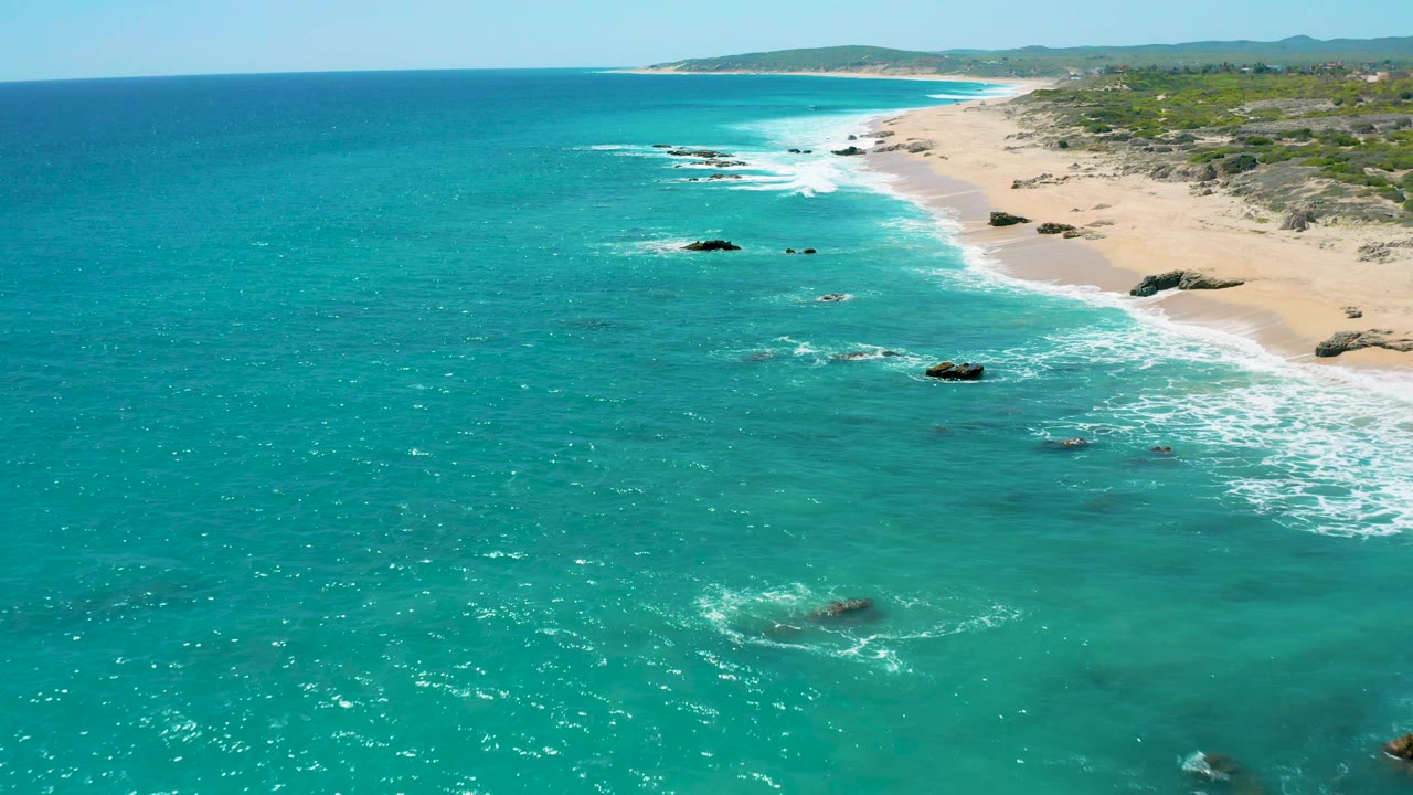 Drone perspective over El Cardoncito, Baja California Sur, showing a long vacant beach curving into the distance, small offshore rocks, bright turquoise water, and no visible people.
