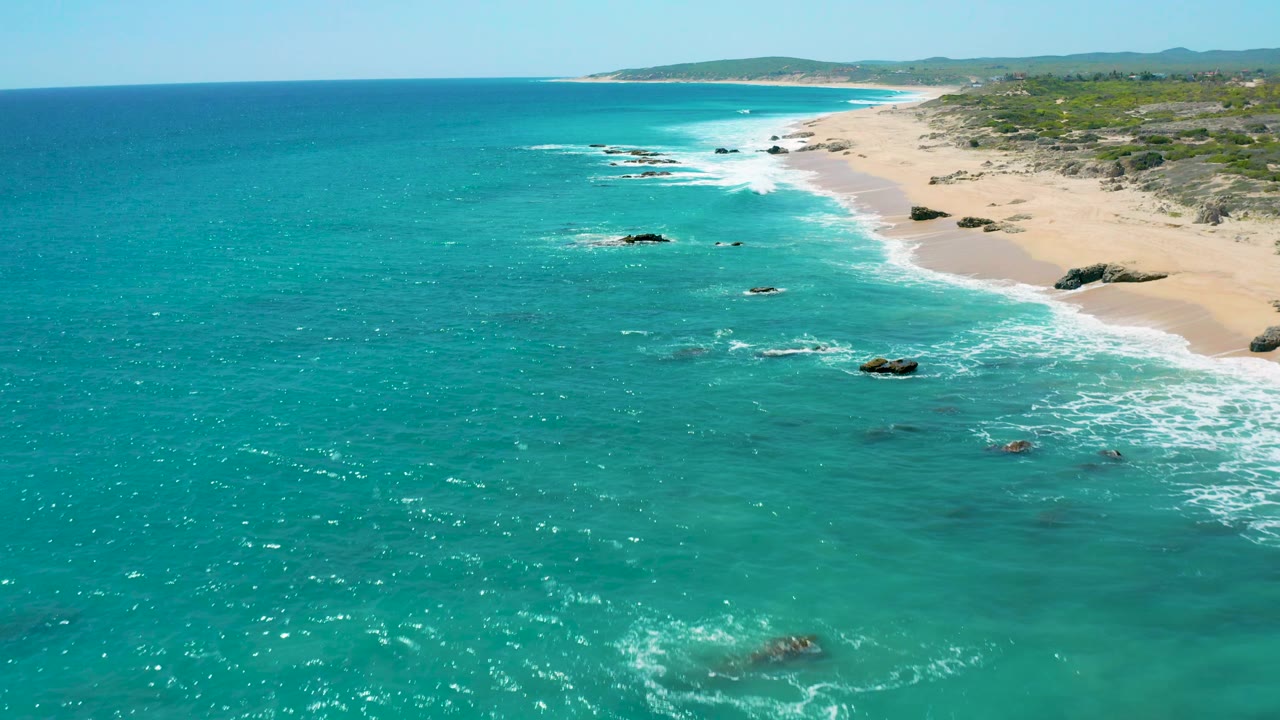 Wide aerial frame of El Cardoncito, Baja California Sur, with open turquoise water on the left, sandy beach and scrubland on the right, scattered rocks near the surf, and no visible people.