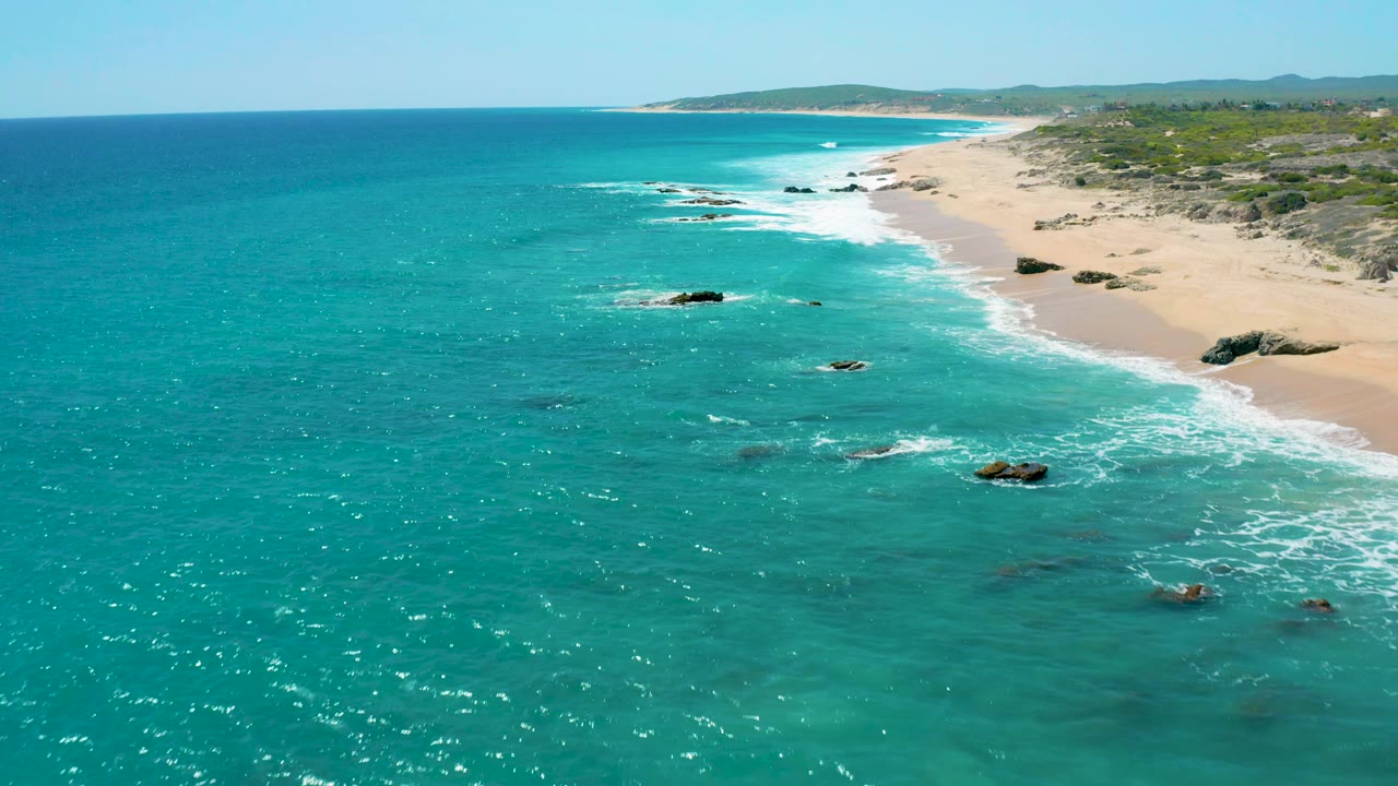 Aerial view at El Cardoncito, Baja California Sur, with turquoise sea in the foreground, foamy waves reaching the sand, dark rocks near shore, low vegetation behind the beach, and no visible people.