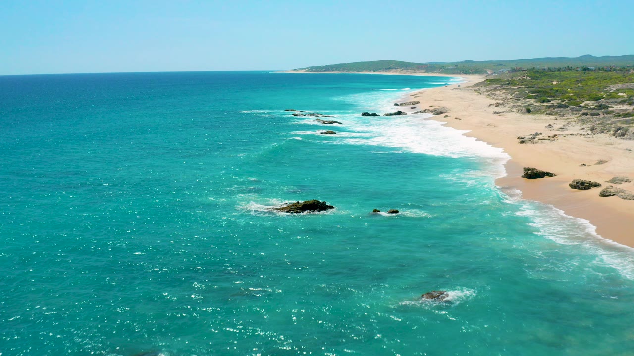 Aerial view over El Cardoncito with turquoise water in the foreground, a pale sandy beach curving along the right side, dark rocks in the shallows, and no visible people.
