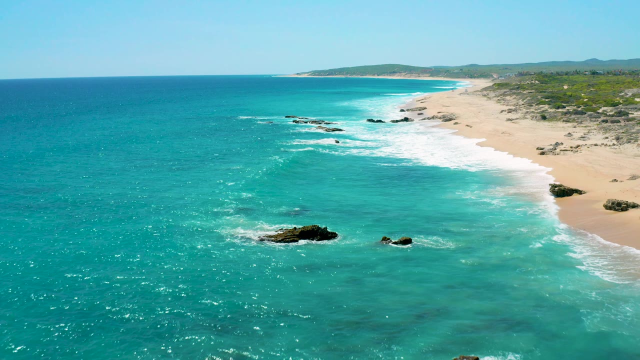 Aerial view over El Cardoncito with a broad field of turquoise ocean, a surf band tracing the sand, rocky outcrops near the lower center, and no visible people.