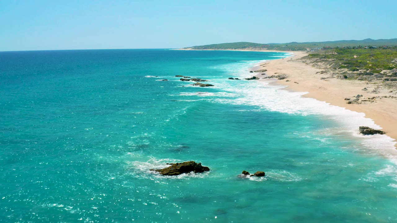 Aerial frame at El Cardoncito with dark rocks near the lower edge, sunlit turquoise shallows, a white wave line along the right-side beach, and no visible people.