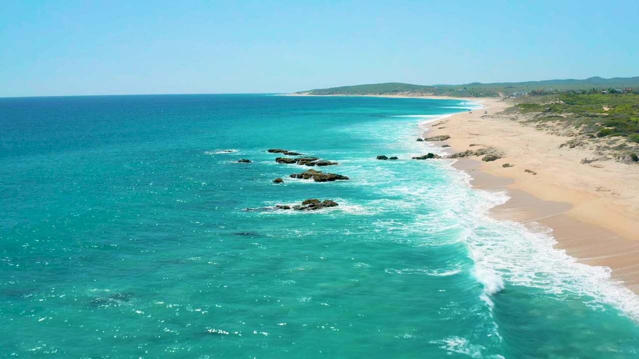 Aerial frame over El Cardoncito showing a long pale beach on the right, turquoise water on the left, small rock groups in the surf zone, a clear horizon, and no visible people.