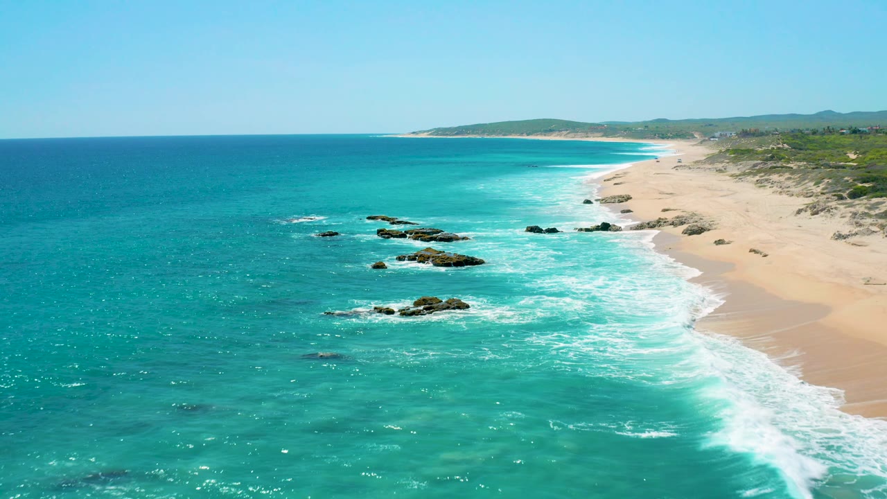 El Cardoncito aerial frame with darker rock clusters more central in the turquoise shallows, foamy surf sweeping toward the right-side beach, distant low hills, and no visible people.