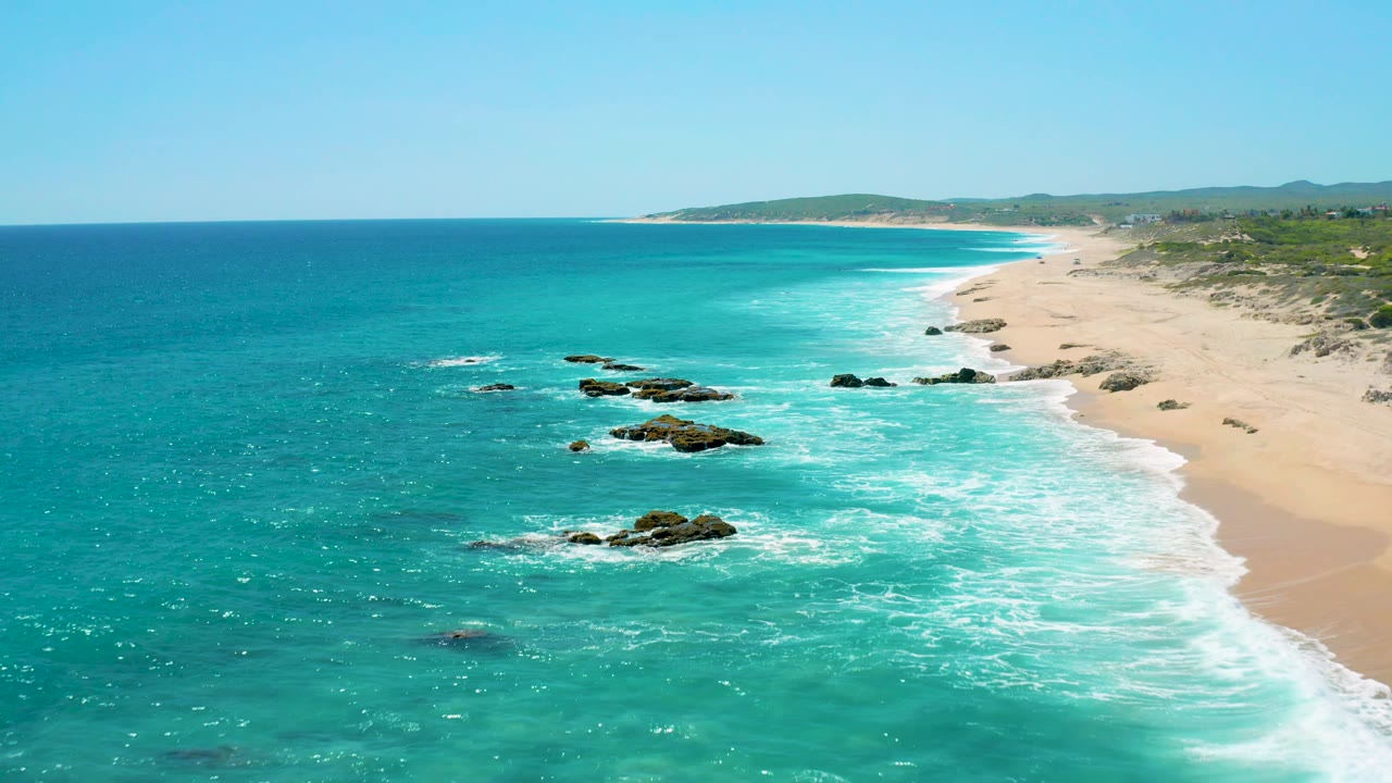 Wide aerial frame over El Cardoncito with a broad span of turquoise ocean, several dark rocks in the surf, a pale beach curving into the distance, and no visible people.