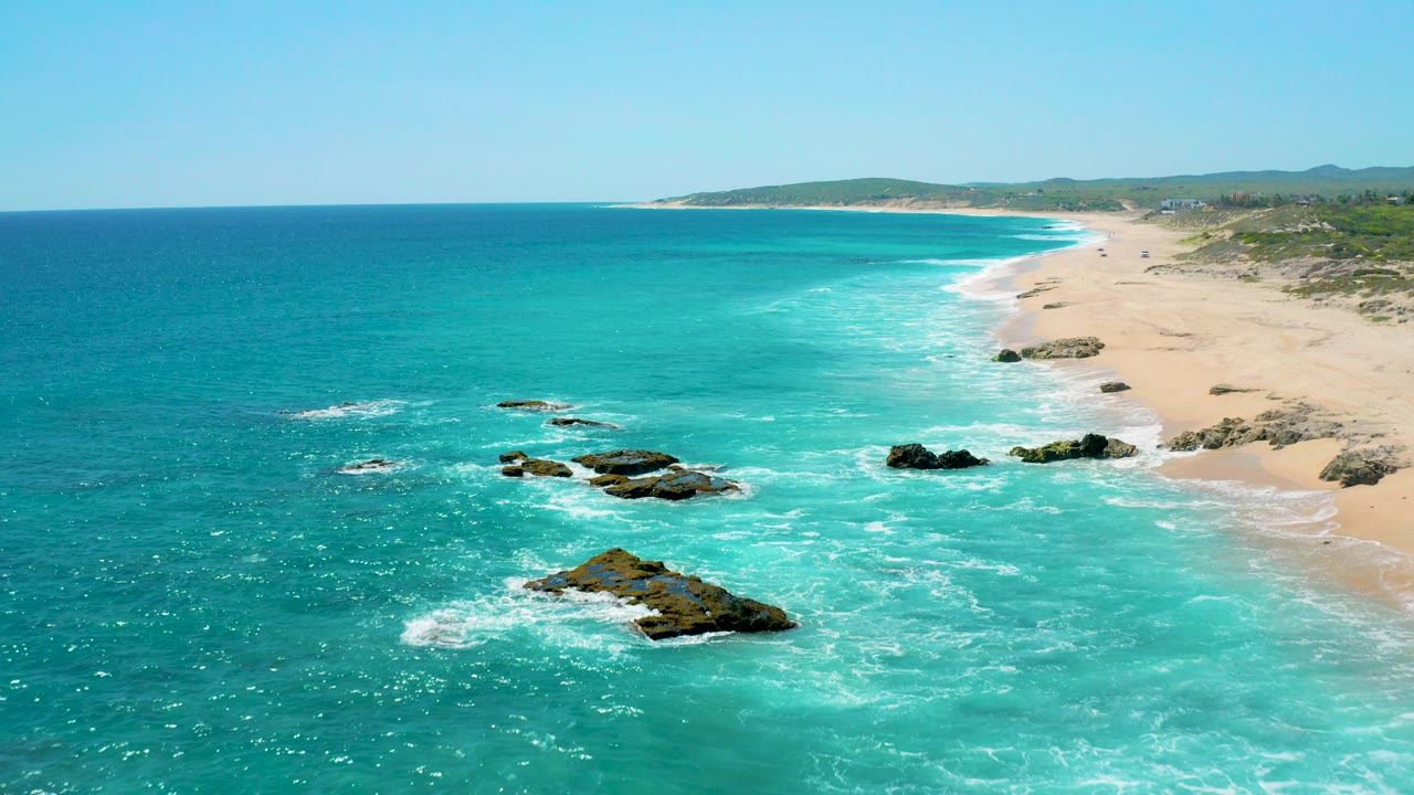 Aerial view over El Cardoncito with turquoise water, white surf around dark rocks, a long sandy beach, low green hills in the distance, and no clearly visible people.