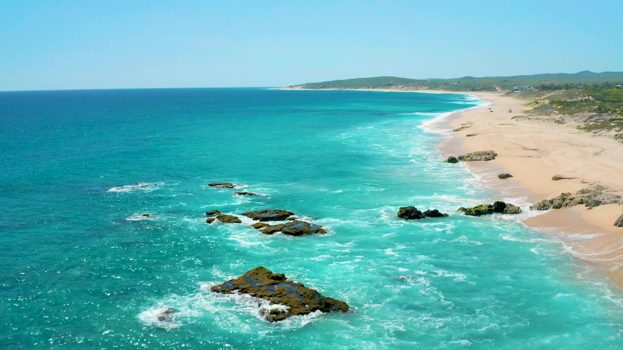 Aerial coastal frame from El Cardoncito with several dark rocks surrounded by white foam, clear turquoise surf, open tan sand on the right, and low hills across the horizon.