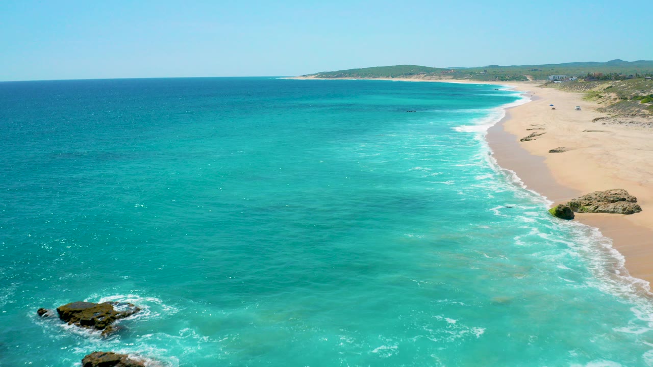 Aerial view over El Cardoncito, Baja California Sur, with turquoise ocean on the left, white surf along the sandy beach on the right, distant low hills, and no visible people.