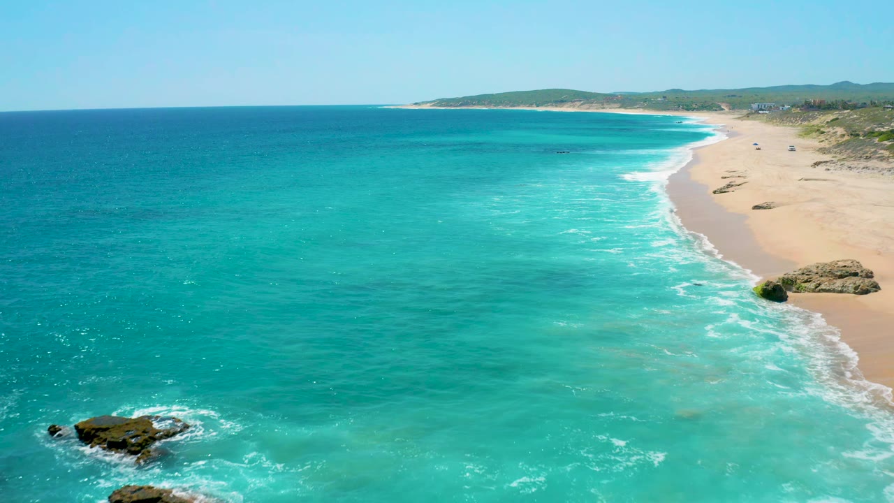 Aerial frame over El Cardoncito showing clear turquoise water across most of the image, a curving sandy beach at right, small dark rocks near the lower edge, and no visible people.