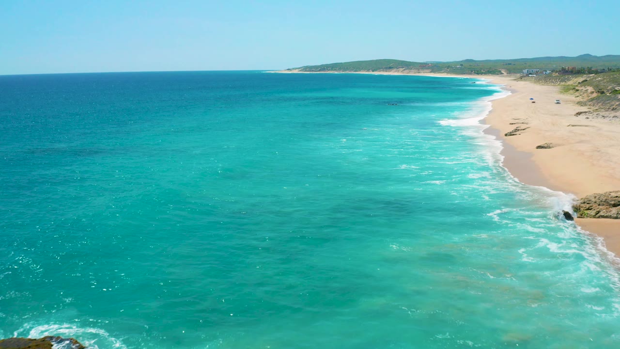Wide aerial view at El Cardoncito with bright teal water in the foreground, pale sand and breaking surf along the right edge, low dunes and hills beyond, and no visible people.