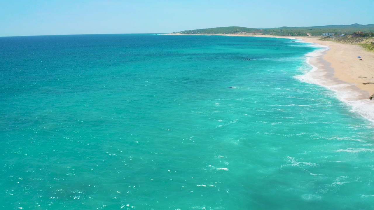 High aerial view over turquoise water and a pale sandy beach at El Cardoncito, Baja California Sur, with low hills on the horizon and no visible people.