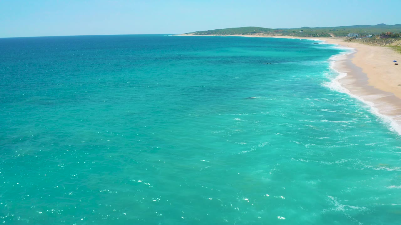 Wide aerial coastline at El Cardoncito showing turquoise shallows, a long sandy beach on the right, low vegetation inland, and no visible people.